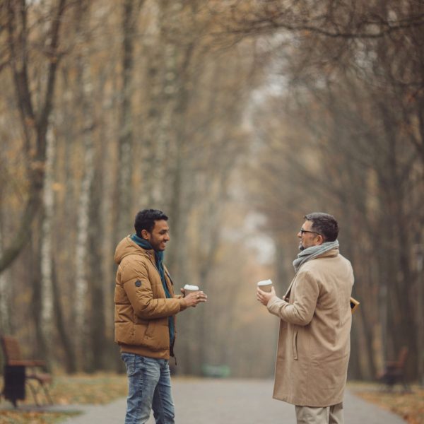 Friends, a senior and a young man walking and talking and drinking coffee together in the autumn park.